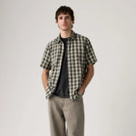 Man wearing a plaid shirt over a black t-shirt with beige pants on a plain background