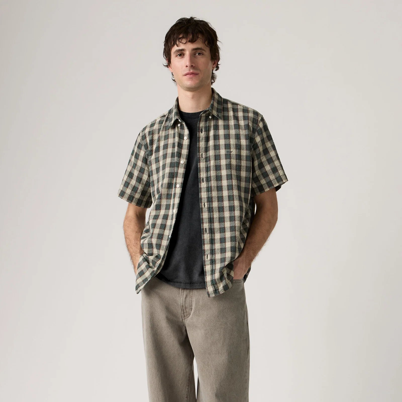 Man wearing a plaid shirt over a black t-shirt with beige pants on a plain background